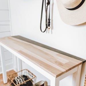 a white wooden console table with a black metal shelf underneath, along with a pair of black rain boots and a white hat hanging on the wall.