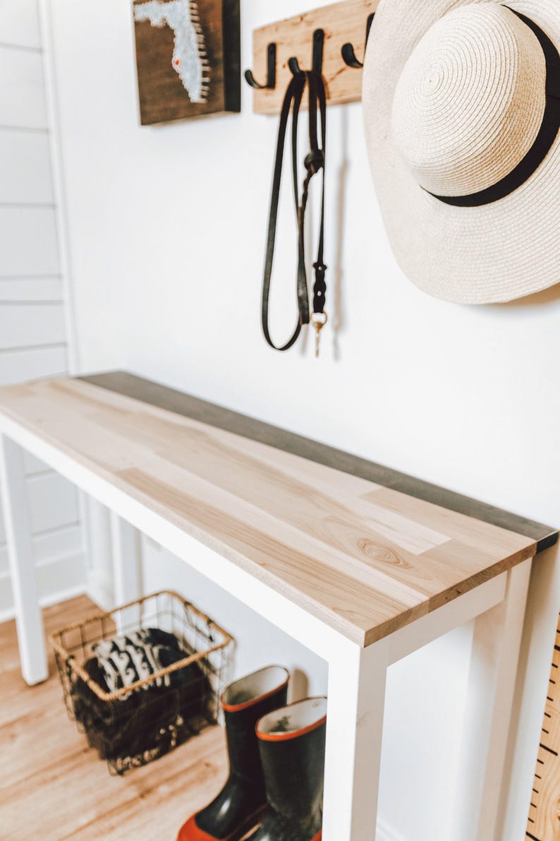 a white wooden console table with a black metal shelf underneath, along with a pair of black rain boots and a white hat hanging on the wall.