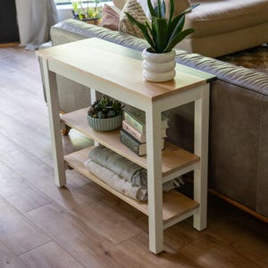 A white side table with a plant and books on it, situated in a living room with a couch and a window.
