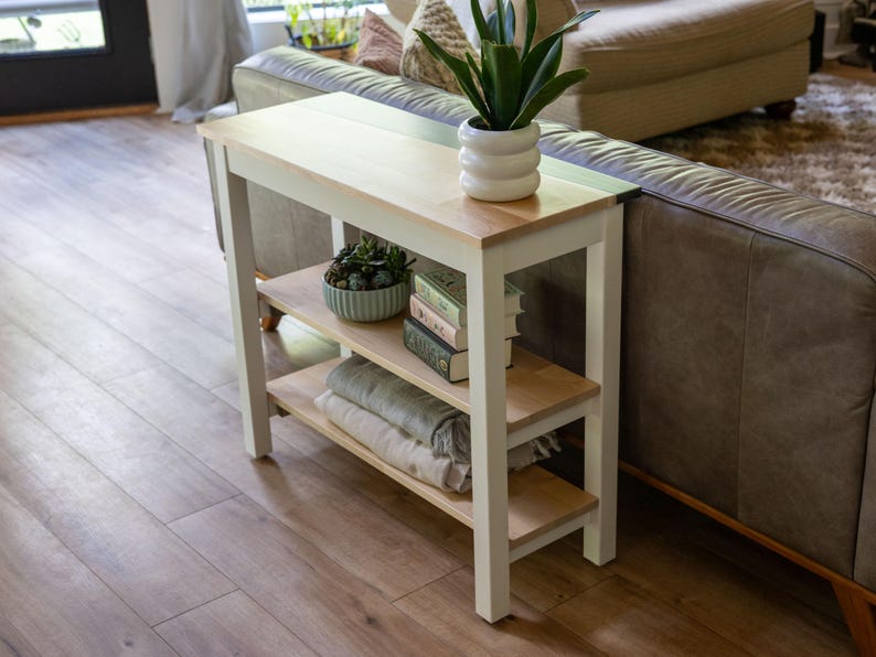 A white side table with a plant and books on it, situated in a living room with a couch and a window.