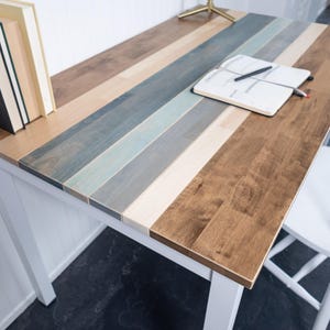 May include: A wooden desk with a multi-colored striped top in shades of brown, blue, and white. The desk has white legs and a white chair is next to it. Books and a notebook with a pen are on the desk.