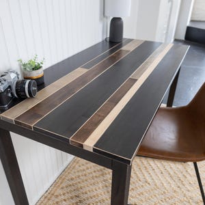 May include: A dark wood desk with a multi-toned planked top. The desk has a black frame and is paired with a brown leather chair. A vintage camera and small potted plant sit on the desk.