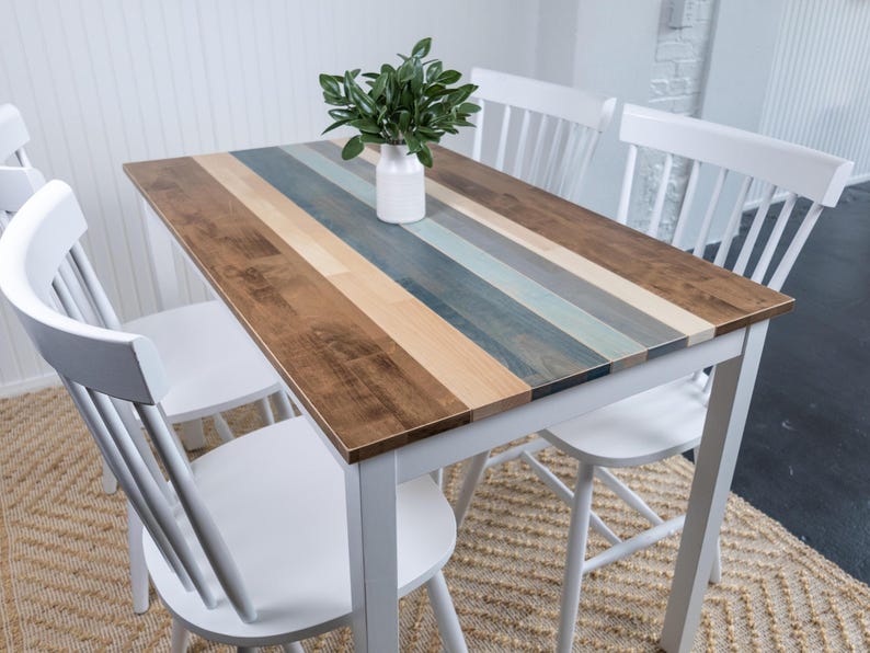 May include: A rectangular wooden table with a multi-coloured striped top, supported by a white frame. The table is surrounded by white chairs. A white vase with green leaves sits on the table. The table is approximately 122 cm long and 76 cm wide.