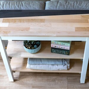 a white wooden side table with a black top, placed on a wooden floor. The table has two shelves, one on the left and one on the right. On the left shelf, there is a blue bowl with a plant inside, and on the right shelf, there are two books stacked on top of each other.