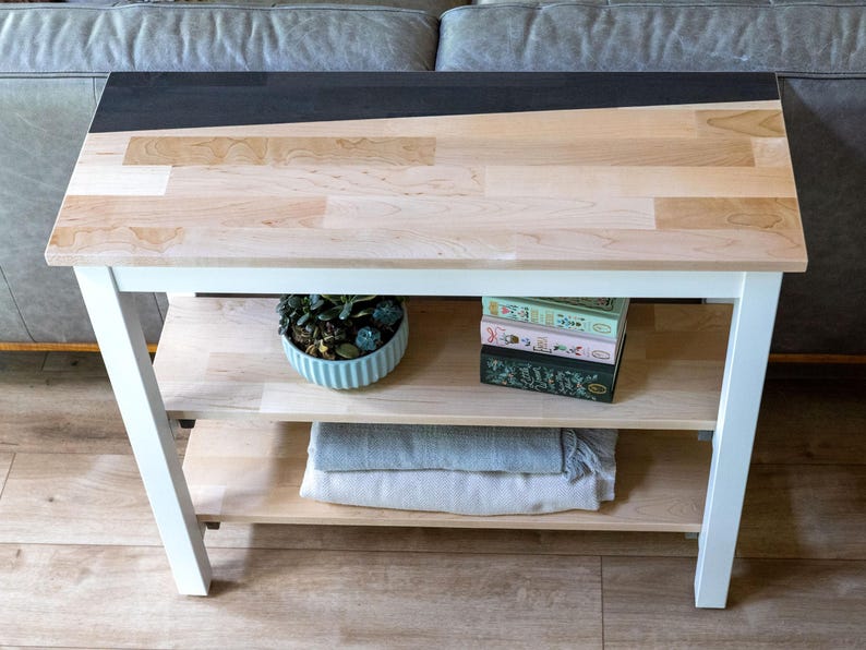 a white wooden side table with a black top, placed on a wooden floor. The table has two shelves, one on the left and one on the right. On the left shelf, there is a blue bowl with a plant inside, and on the right shelf, there are two books stacked on top of each other.