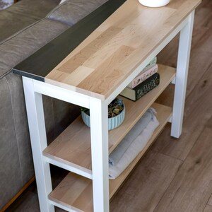 a modern, minimalist living room with a white side table and a gray couch. The table has a wooden top and metal frame, and it has two shelves for storage. The couch is positioned next to the table, creating a cozy and functional seating area.