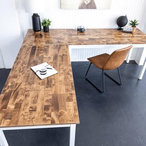 May include: An L-shaped wooden desk with a light brown finish and white legs. A brown leather chair with black legs sits nearby. The desk is accessorized with a black vase, a small plant, and a vintage camera.