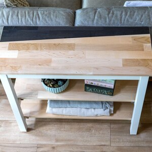 a wooden side table with a black top, placed on a wooden floor in front of a gray couch. The table has two shelves, one on the bottom and one on the top, and it has a few items on it, including a green plant, a blue bowl, and a stack of books.