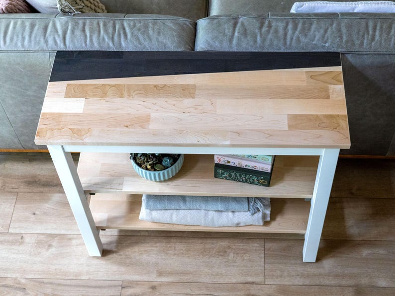 a wooden side table with a black top, placed on a wooden floor in front of a gray couch. The table has two shelves, one on the bottom and one on the top, and it has a few items on it, including a green plant, a blue bowl, and a stack of books.