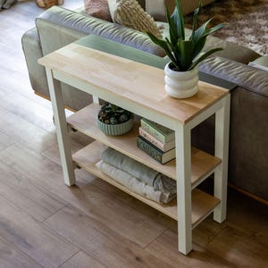 a cozy living room with a wooden side table and a comfortable couch. The table has a shelf on the top and a shelf below, providing storage and display space. On the table, there is a potted plant, a bowl, and a stack of books. The couch is positioned against the wall, and the floor is made of wood.
