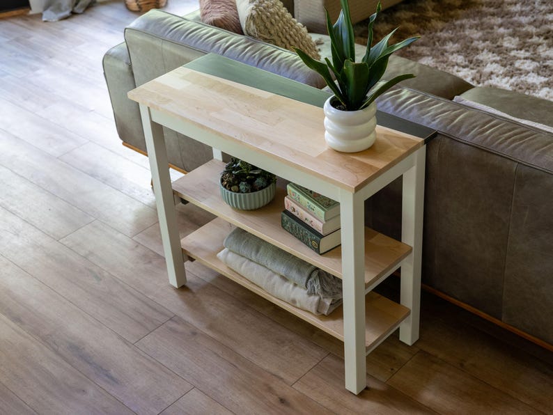 a cozy living room with a wooden side table and a comfortable couch. The table has a shelf on the top and a shelf below, providing storage and display space. On the table, there is a potted plant, a bowl, and a stack of books. The couch is positioned against the wall, and the floor is made of wood.