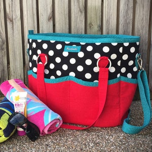 May include: A black and white polka dot beach tote bag with teal trim and red lining. The bag has two handles and a shoulder strap. The bag is sitting on a gravel surface with a rolled up towel, sunscreen, and sunglasses.