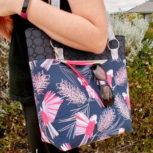 May include: A tote bag featuring a dark blue fabric with a pink and white galah cockatoo and floral pattern, a black upper panel with a circular geometric print, silver metal rings, and a pink tassel. A pair of black sunglasses hangs from the strap.