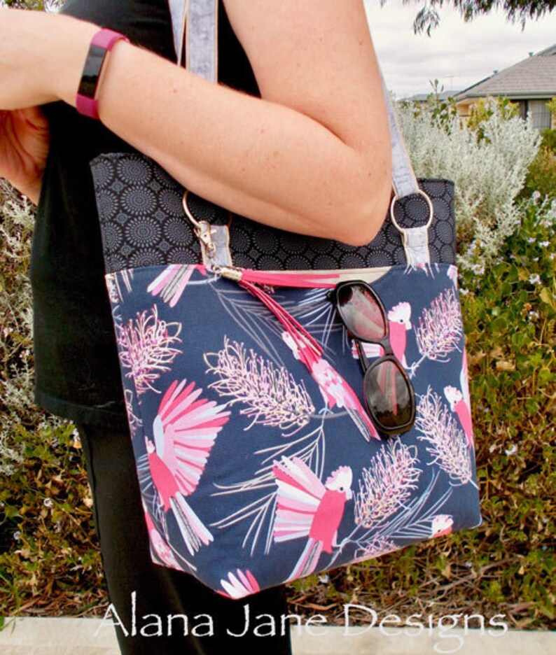 May include: A tote bag featuring a dark blue fabric with a pink and white galah cockatoo and floral pattern, a black upper panel with a circular geometric print, silver metal rings, and a pink tassel. A pair of black sunglasses hangs from the strap.