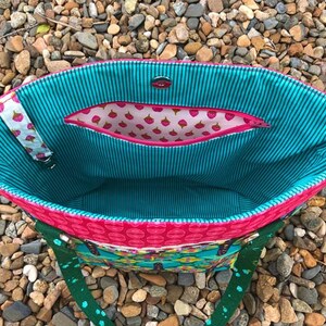 May include: A tote bag interior featuring a teal and black pinstriped lining, a pink zippered pocket with a strawberry print, a magnetic snap closure, and a white fabric strap with a strawberry pattern attached to the side of the bag.