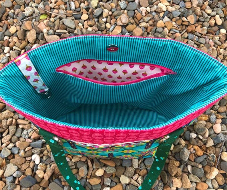 May include: A tote bag interior featuring a teal and black pinstriped lining, a pink zippered pocket with a strawberry print, a magnetic snap closure, and a white fabric strap with a strawberry pattern attached to the side of the bag.