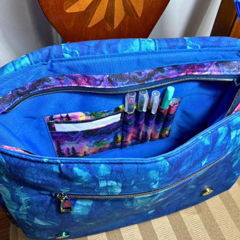 May include: A blue canvas tote bag with a zipper closure and a colourful patterned lining. The bag has a zippered pocket on the front and a small pocket on the inside. The inside pocket has a white card slot and several pen slots.