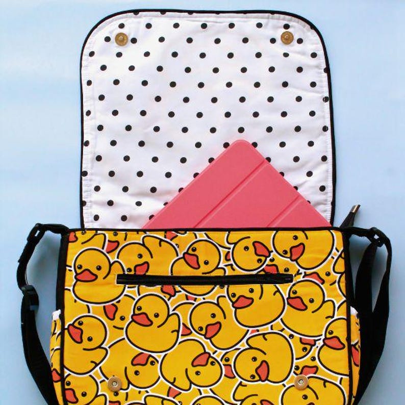 May include: A black messenger bag with a white polka dot lining and a zip closure. The bag is decorated with a repeating pattern of yellow rubber ducks. A pink tablet computer is inside the bag.