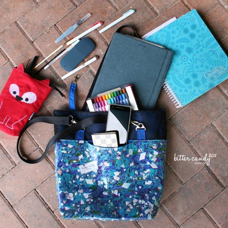 May include: A blue and black tote bag with a mosaic pattern, containing a smartphone, a small wallet, and a set of markers. Nearby are a red fox-shaped pencil case, a dark gray tablet, a light blue spiral-bound planner, and several pens.