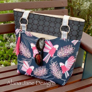 May include: A tote bag featuring a dark blue fabric with a pink cockatoo and floral print, a black patterned upper section, light-colored straps with silver rings, a pink tassel, and a pair of black sunglasses tucked into the front pocket.