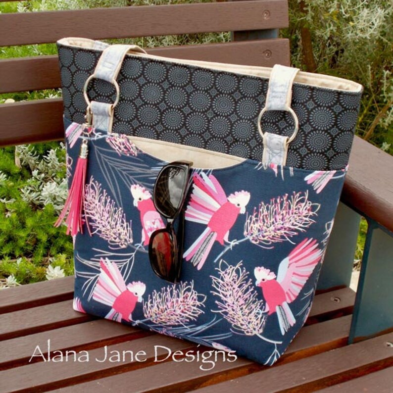 May include: A tote bag featuring a dark blue fabric with a pink cockatoo and floral print, a black patterned upper section, light-colored straps with silver rings, a pink tassel, and a pair of black sunglasses tucked into the front pocket.