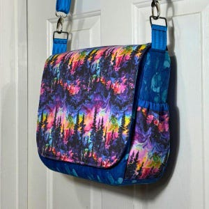 May include: A blue and purple messenger bag with a colourful forest scene print. The bag has a flap closure and a zipped pocket on the front.