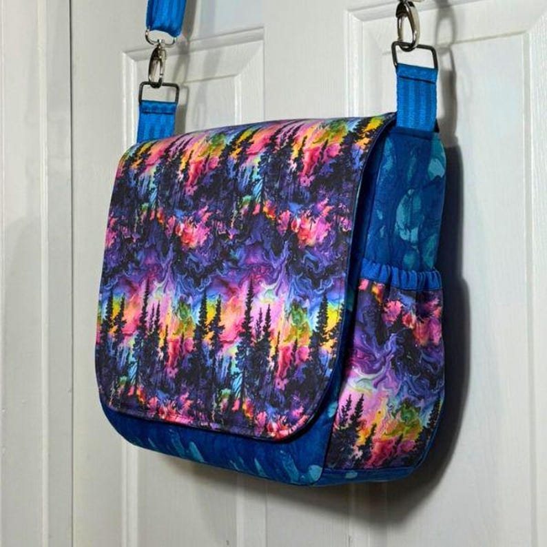 May include: A blue and purple messenger bag with a colourful forest scene print. The bag has a flap closure and a zipped pocket on the front.