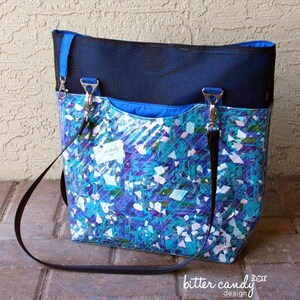 May include: A tote bag featuring a top section of black mesh and a lower section with a vibrant blue, purple, and white geometric pattern. The bag includes black straps attached with silver metal hardware and a small white tag with text.