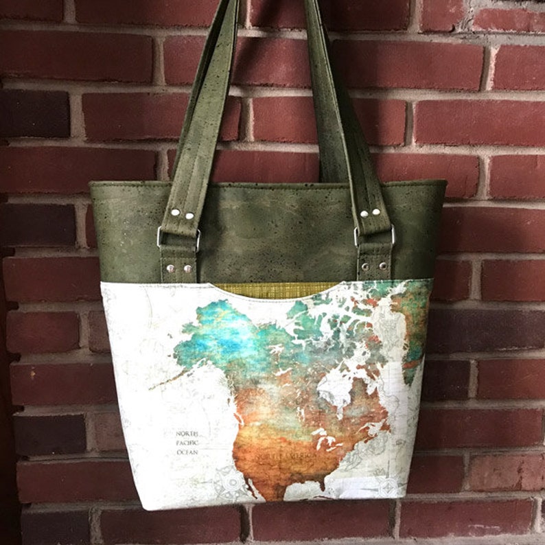 May include: A tote bag featuring a top section made of olive green cork material and a bottom section printed with a colorful map of North America. The bag includes two matching olive green cork shoulder straps attached with silver metal rivets.
