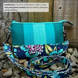 May include: A turquoise and blue fabric clutch with a floral print strap. The clutch measures 9 inches wide by 6 inches high by 0.5 inches deep. "Round It Up Clutch" is written in white text on a brown background.
