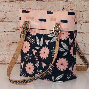 May include: A vertical tote bag featuring a dark navy blue lower section with a pink floral and leaf pattern. The upper section is light pink with dark navy brushstroke accents. It includes two cork-textured shoulder straps attached with gold rings.