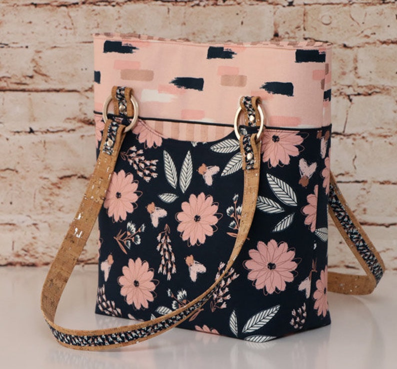 May include: A vertical tote bag featuring a dark navy blue lower section with a pink floral and leaf pattern. The upper section is light pink with dark navy brushstroke accents. It includes two cork-textured shoulder straps attached with gold rings.
