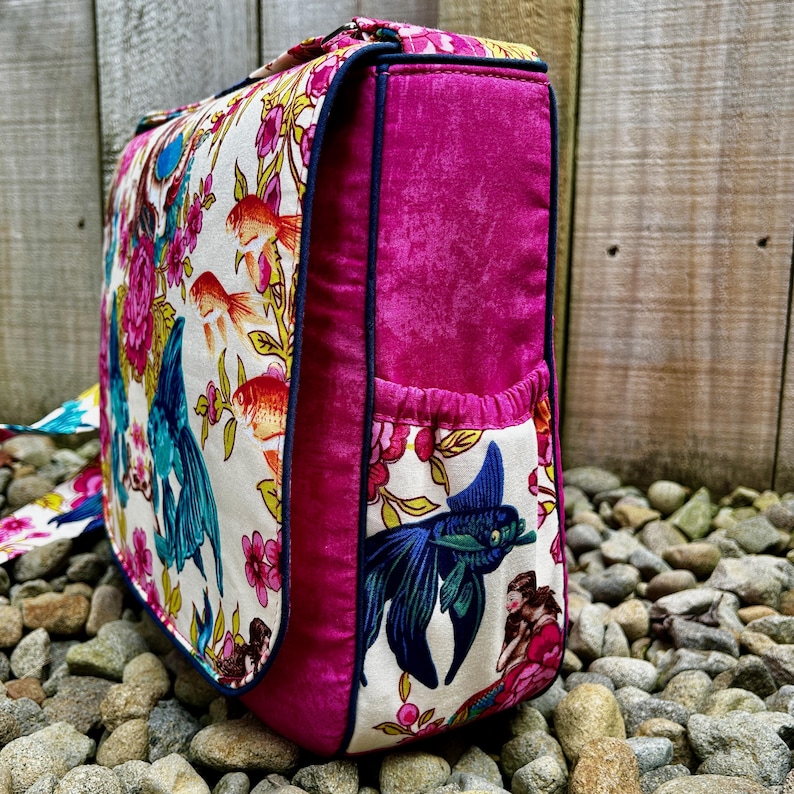 May include: A pink and white fabric messenger bag with a floral and fish design. The bag has a flap closure and a small pocket on the front.