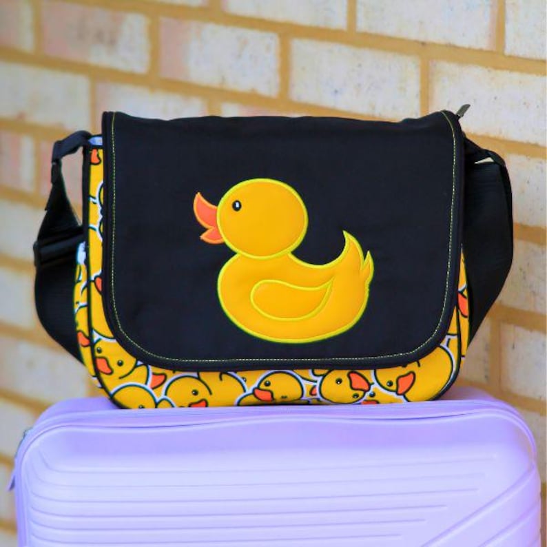 May include: A black messenger bag featuring a yellow rubber ducky design. The bag has a flap closure and a shoulder strap. The bag is decorated with a pattern of yellow rubber duckies.