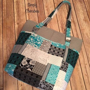 May include: A tote bag featuring a patchwork design with teal, black, white, and gray fabric squares. The patterns include polka dots, floral swirls, and geometric shapes. The text Simply Cherabea is printed in black on the upper left side.