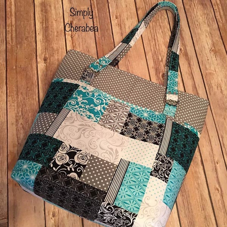 May include: A tote bag featuring a patchwork design with teal, black, white, and gray fabric squares. The patterns include polka dots, floral swirls, and geometric shapes. The text Simply Cherabea is printed in black on the upper left side.