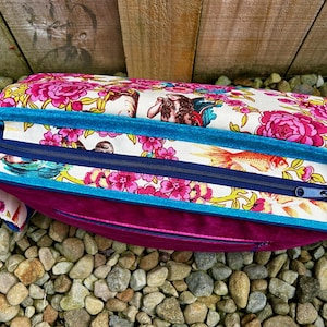 May include: A pink and white floral fabric bag with a teal blue trim and a zipper closure. The bag is sitting on a bed of small stones.