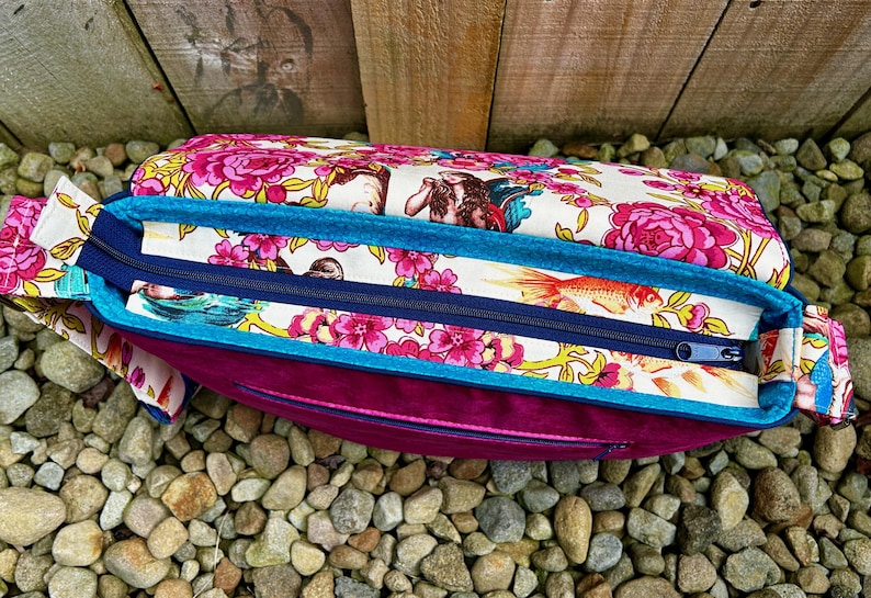 May include: A pink and white floral fabric bag with a teal blue trim and a zipper closure. The bag is sitting on a bed of small stones.