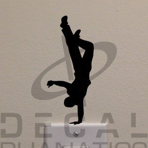 May include: A black silhouette of a person doing a handstand on a white light switch cover. The switch cover has the text "Decal Phanatics" printed on it.