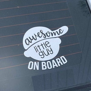 May include: White vinyl sticker with the text "awesome little guy on board" in a black circle with a baseball cap on top.