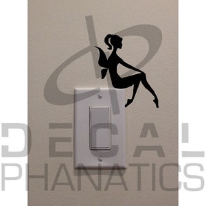 Puede incluir: Un interruptor de luz blanco con una silueta negra de un hada sentada encima. El hada tiene alas y lleva un vestido.