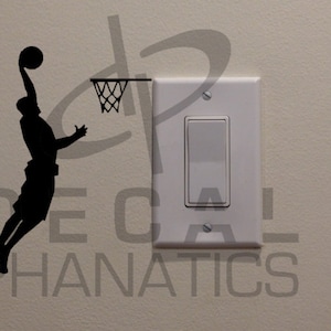 Könnte beinhalten: Schwarze Silhouette eines Basketballspielers in der Luft, kurz davor, den Ball zu dunken. Der Spieler befindet sich neben einem Basketballkorb und einem weißen Lichtschalter an einer beigefarbenen Wand. Die Worte "DECAL PHANATICS" sind sichtbar.