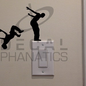 May include: Two black silhouette figures of people performing acrobatic stunts. The figure on the left is upside down, while the figure on the right is standing on a light switch cover. The text "DECAL PHANATICS" is visible below the figures.