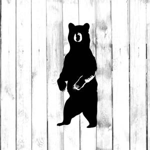 May include: Black silhouette of a bear standing on its hind legs with its paws raised in front of it. The bear is facing forward with its head tilted slightly down. The background is a white wooden fence.