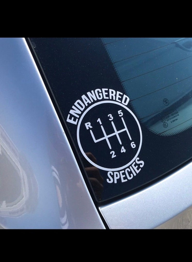 May include: A white vinyl sticker on a car window with a black circle and the text "ENDANGERED SPECIES" around it. Inside the circle is a white gear shift pattern with the letters "R" and numbers "1", "2", "3", "4", "5", and "6".