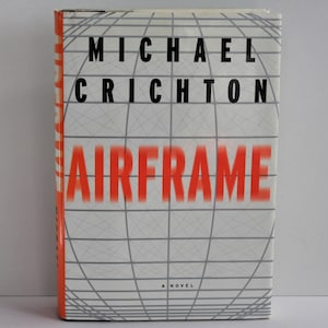 Può includere: Una copertina di libro bianca con un motivo a griglia rosso e bianco. Il titolo "AIRFRAME" è in lettere rosse. Il nome dell'autore, "MICHAEL CRICHTON", è in lettere nere. Il libro è un romanzo.
