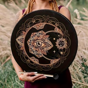 Puede incluir: Un tambor de marco redondo negro con una ilustración dorada detallada de un ojo central rodeado de intrincados patrones florales, una luna creciente y un pequeño sol. Un mazo recto y delgado se sostiene horizontalmente frente al tambor.