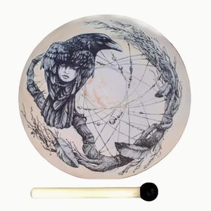 May include: A circular drum featuring a detailed black and white illustration of a raven perched on a branch, with a human face integrated into the bird's plumage, surrounded by a webbed dreamcatcher design and feathers. A wooden mallet is included.