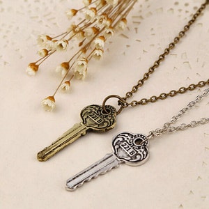 May include: Two silver key necklaces with antique-style key pendants. The keys have a heart-shaped head with the number "218" engraved on them. The necklaces are on a white background with dried flowers.