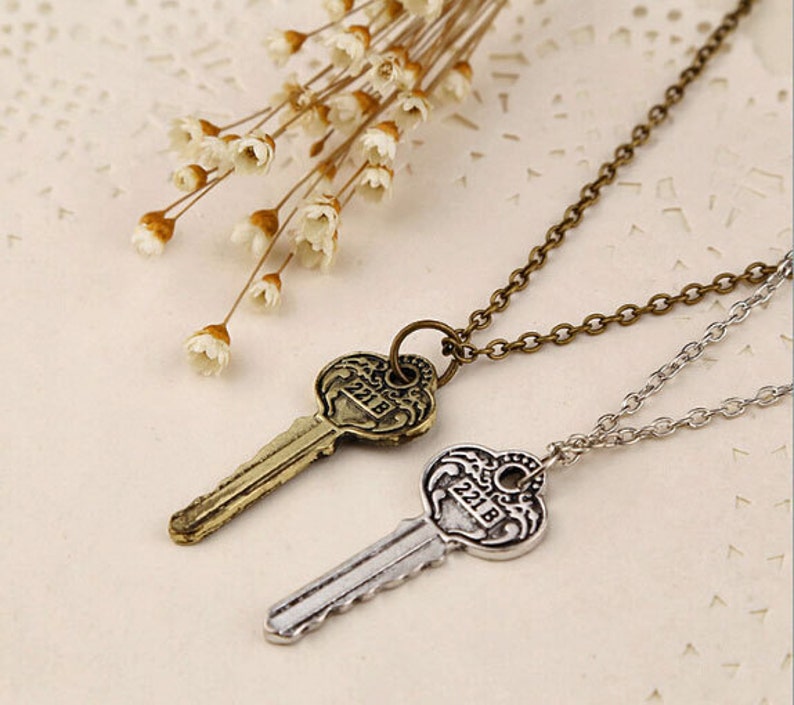 May include: Two silver key necklaces with antique-style key pendants. The keys have a heart-shaped head with the number "218" engraved on them. The necklaces are on a white background with dried flowers.